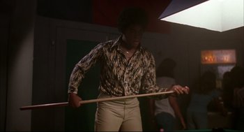 Movie still from “Dazed and Confused” (1993), directed by Richard Linklater – A man holding a wooden stick while standing in a darkened room; Medium shot, Low angle
