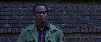 Movie still from “Dead Presidents” (1995), directed by Albert Hughes – A man in a green jacket and striped shirt standing in front of a brick wall; Close Up shot, Over the shoulder angle