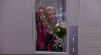 Movie still from “Death Becomes Her” (1992), directed by Robert Zemeckis – A woman holding a wine glass and a bouquet of flowers in front of a mirror; Medium shot, Over the shoulder angle