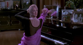 Movie still from “Death Becomes Her” (1992), directed by Robert Zemeckis – A woman in a purple dress standing in front of a piano; Medium shot, Over the shoulder angle