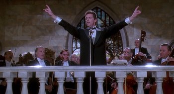 Movie still from “Death Becomes Her” (1992), directed by Robert Zemeckis – A man in a suit and bow tie holding a microphone; Medium shot, Low angle