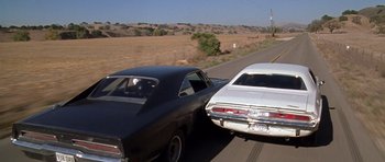 Movie still from “Death Proof” (2007), directed by Quentin Tarantino – A couple of cars driving down a road next to a field; Wide shot, High angle