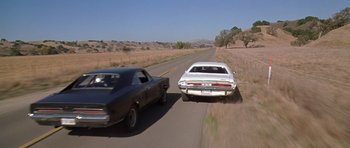 Movie still from “Death Proof” (2007), directed by Quentin Tarantino – Two cars are driving down a road near a grassy field; Wide shot, High angle