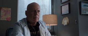 Movie still from “Death Wish” (2018), directed by Eli Roth – A bald man wearing a lab coat sitting in front of a lamp; Close Up shot, Low angle