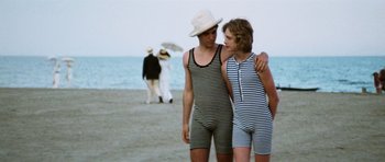 Movie still from “Death in Venice” (1971), directed by Luchino Visconti – A man and a woman standing next to each other on the beach; Medium shot, Low angle