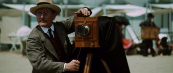 Movie still from “Death in Venice” (1971), directed by Luchino Visconti – A man holding an old fashioned camera on top of a tripod; Close Up shot, Low angle