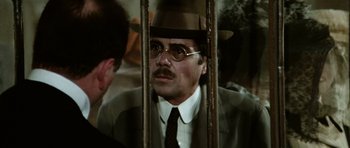 Movie still from “Death in Venice” (1971), directed by Luchino Visconti – A man in a suit and a hat is sitting behind bars; Close Up shot, Over the shoulder angle