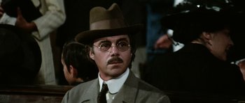Movie still from “Death in Venice” (1971), directed by Luchino Visconti – A man in a suit and a hat is wearing glasses; Close Up shot, Low angle