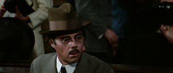 Movie still from “Death in Venice” (1971), directed by Luchino Visconti – A man in a hat and glasses sitting in front of other men; Close Up shot, Low angle