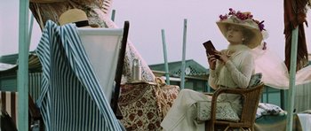 Movie still from “Death in Venice” (1971), directed by Luchino Visconti – A woman sitting on a chair reading a book while holding a cell phone to her ear; Medium shot, Low angle