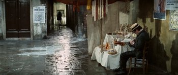 Movie still from “Death in Venice” (1971), directed by Luchino Visconti – A person walking down a street near a table with a lot of stuff on it; Wide shot, High angle