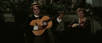 Movie still from “Death in Venice” (1971), directed by Luchino Visconti – A man in a suit and hat holding a guitar; Medium shot, High angle