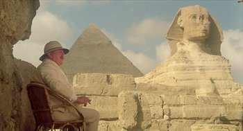 Movie still from “Death on the Nile” (1978), directed by John Guillermin – An older man sitting in a chair in front of a pyramid; Wide shot, Low angle