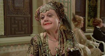 Movie still from “Death on the Nile” (1978), directed by John Guillermin – An older woman wearing a headdress and holding a glass of red wine; Close Up shot, Over the shoulder angle