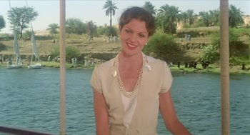 Movie still from “Death on the Nile” (1978), directed by John Guillermin – A woman wearing a white shirt and a pearl necklace standing in front of a body of water; Medium shot, Low angle