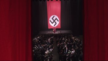 Movie still from “Death to Smoochy” (2002), directed by Danny DeVito – A crowd of people sitting in front of a stage with a man standing on it; Extreme Wide shot, High angle