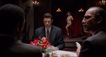 Movie still from “Deep Cover” (1992), directed by Bill Duke – A man sitting at a dinner table with a woman standing in the background; Medium shot, Over the shoulder angle