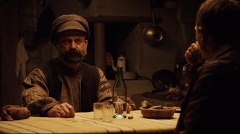 Movie still from “Defiance” (2008), directed by Edward Zwick – A man sitting at a table with a bottle of wine; Medium shot, Over the shoulder angle