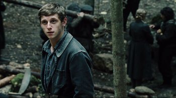 Movie still from “Defiance” (2008), directed by Edward Zwick – A young man standing next to a tree in the woods; Medium shot, Over the shoulder angle