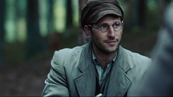 Movie still from “Defiance” (2008), directed by Edward Zwick – A man wearing glasses and a hat is sitting down; Close Up shot, Over the shoulder angle