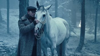 Movie still from “Defiance” (2008), directed by Edward Zwick – A man standing next to a white horse in the snow; Medium shot, Low angle
