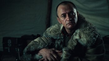 Movie still from “Defiance” (2008), directed by Edward Zwick – A man wearing a fur coat sitting in front of a tent; Close Up shot, Over the shoulder angle