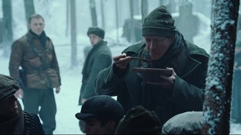 Movie still from “Defiance” (2008), directed by Edward Zwick – A man eating something while standing in the snow; Medium shot, Over the shoulder angle