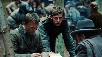 Movie still from “Defiance” (2008), directed by Edward Zwick – Two men sitting next to each other in a crowd of people; Medium shot, Over the shoulder angle