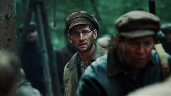Movie still from “Defiance” (2008), directed by Edward Zwick – A man wearing glasses and a hat is standing in the woods; Close Up shot, Over the shoulder angle