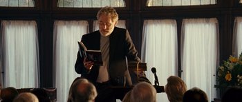 Movie still from “Definitely, Maybe” (2008), directed by Adam Brooks – A man reading a book in front of a microphone; Medium shot, Over the shoulder angle