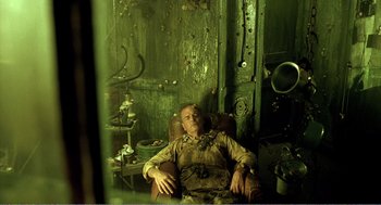 Movie still from “Delicatessen” (1991), directed by Marc Caro – A man sitting in a chair in front of a green wall; Wide shot, High angle