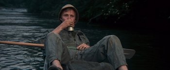 Movie still from “Deliverance” (1972), directed by John Boorman – A man sitting on the ground holding a drink; Medium shot, High angle