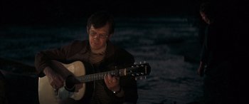 Movie still from “Deliverance” (1972), directed by John Boorman – A man holding a guitar while standing in front of a body of water at night; Medium shot, Over the shoulder angle