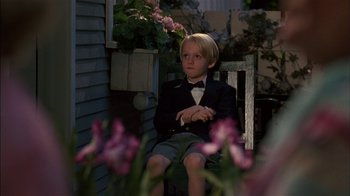 Movie still from “Dennis the Menace” (1993), directed by Nick Castle – A young boy sitting on a chair in front of flowers; Medium shot, Over the shoulder angle