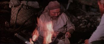 Movie still from “Dersu Uzala” (1975), directed by Akira Kurosawa – An old man wearing a hat and holding a knife in front of a fire; Medium shot, High angle