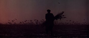 Movie still from “Dersu Uzala” (1975), directed by Akira Kurosawa – A man standing in a field holding a bunch of plants; Wide shot, Low angle