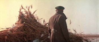 Movie still from “Dersu Uzala” (1975), directed by Akira Kurosawa – A man wearing a coat and hat standing in front of a pile of grass; Medium shot, Low angle