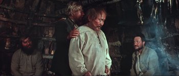Movie still from “Dersu Uzala” (1975), directed by Akira Kurosawa – A man standing next to another man in front of a wooden wall; Medium shot, High angle