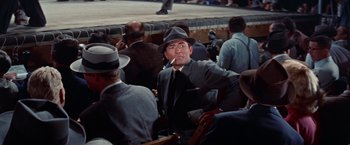 Movie still from “Designing Woman” (1957), directed by Vincente Minnelli – A man smoking a cigarette while sitting in a crowd of people; Medium shot, Low angle
