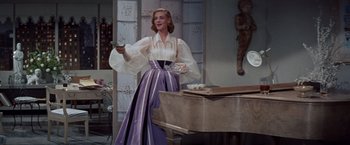 Movie still from “Designing Woman” (1957), directed by Vincente Minnelli – A woman in a white blouse and purple skirt holding a remote control; Medium shot, Low angle