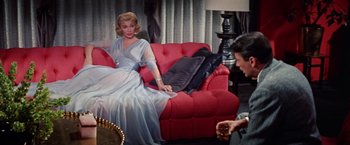 Movie still from “Designing Woman” (1957), directed by Vincente Minnelli – A woman sitting on top of a red couch next to a man; Medium shot, High angle