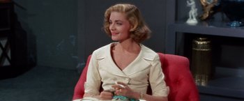 Movie still from “Designing Woman” (1957), directed by Vincente Minnelli – A woman sitting on top of a red chair smoking a cigarette; Medium shot, Low angle