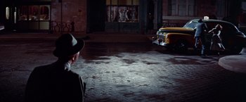 Movie still from “Designing Woman” (1957), directed by Vincente Minnelli – A man wearing a hat and a suit jacket standing in front of a building; Wide shot, Over the shoulder angle