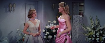 Movie still from “Designing Woman” (1957), directed by Vincente Minnelli – Two women in formal wear talking to each other in front of flowers; Medium shot, Over the shoulder angle