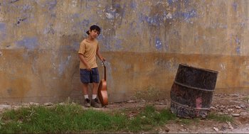 Movie still from “Desperado” (1995), directed by Robert Rodriguez – A young man holding a guitar standing next to a wall; Wide shot, Low angle