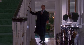 Movie still from “Desperate Hours” (1990), directed by Michael Cimino – A man in a long coat and tie standing in front of a staircase; Wide shot, Low angle