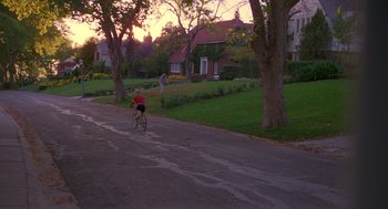 Movie still from “Desperate Hours” (1990), directed by Michael Cimino – A person riding a bike down a street near some trees; Extreme Wide shot, High angle