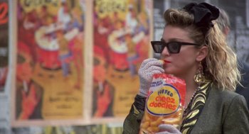 Movie still from “Desperately Seeking Susan” (1985), directed by Susan Seidelman – A woman holding a bag of cheetos while wearing white gloves; Close Up shot, Over the shoulder angle