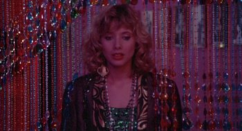 Movie still from “Desperately Seeking Susan” (1985), directed by Susan Seidelman – A woman in a black and gold dress standing in front of beads; Close Up shot, Over the shoulder angle