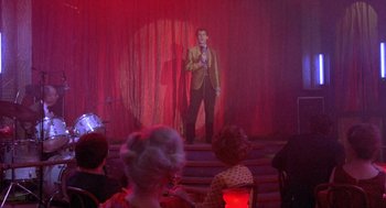 Movie still from “Desperately Seeking Susan” (1985), directed by Susan Seidelman – A man standing on steps in front of a crowd; Wide shot, Over the shoulder angle